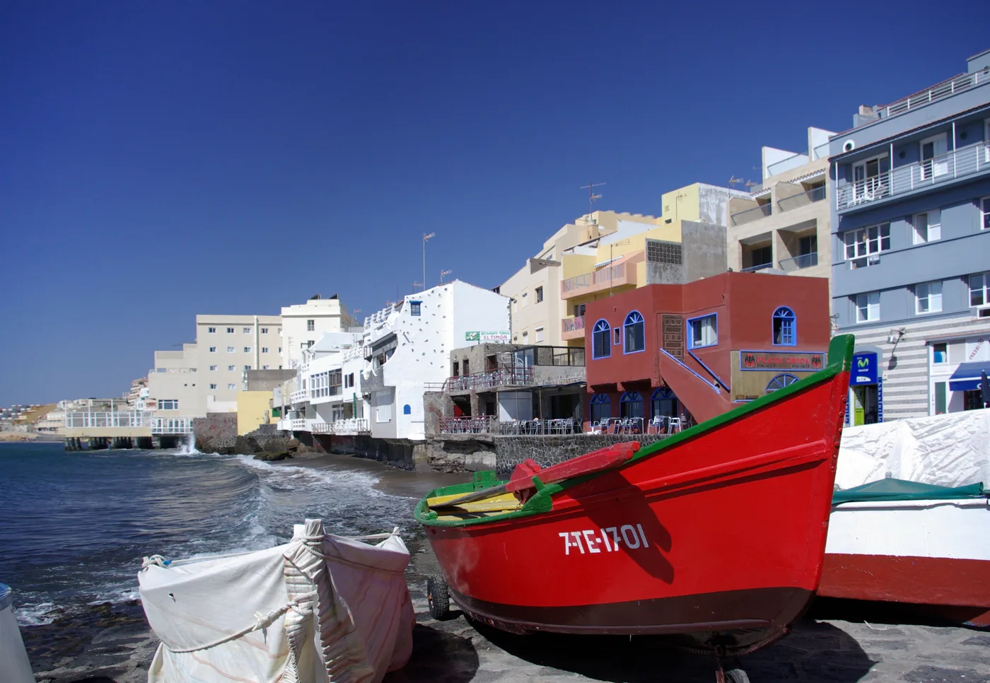 A red fishing boat drawn up on the rocks below the seafront buildings of El Médano, Tenerife.
